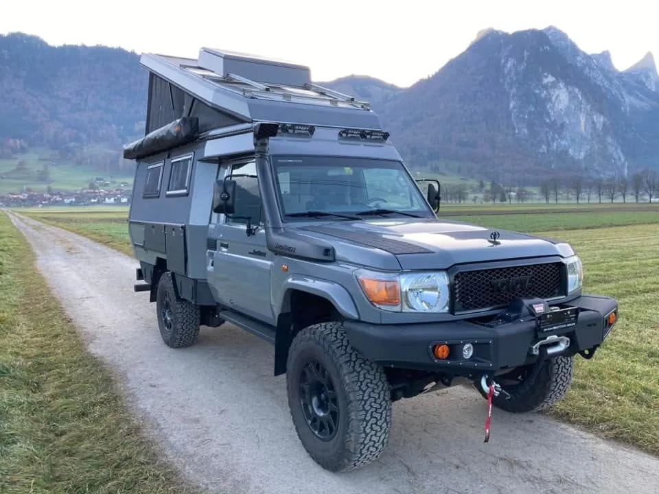 Dark grey Land Cruiser 79 pop-top camper on an Alpine gravel path with dramatic mountain backdrop, showing a premium expedition conversion in a stunning environment.