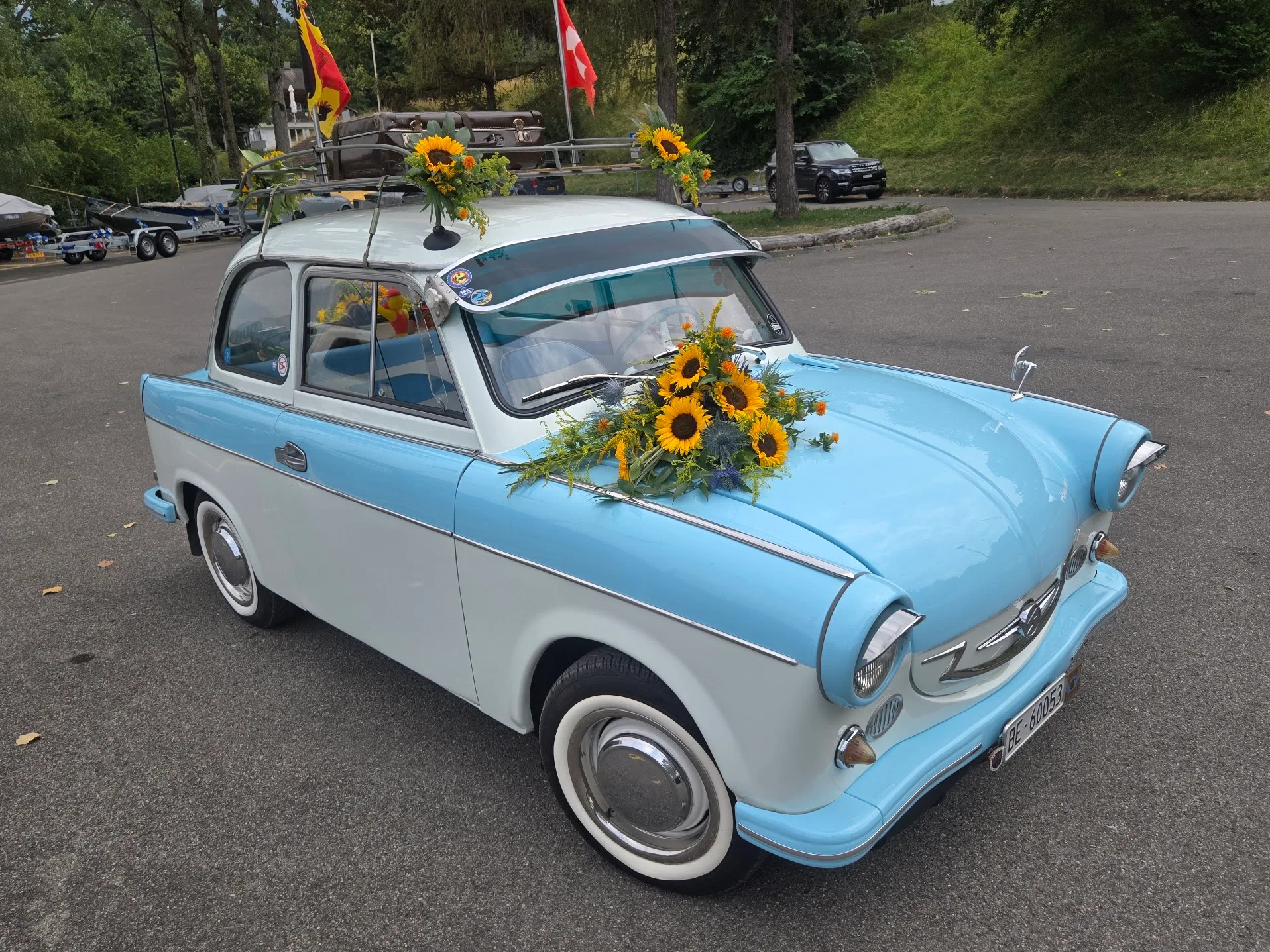A man proudly stands beside a vintage light-blue classic car at an outdoor event.