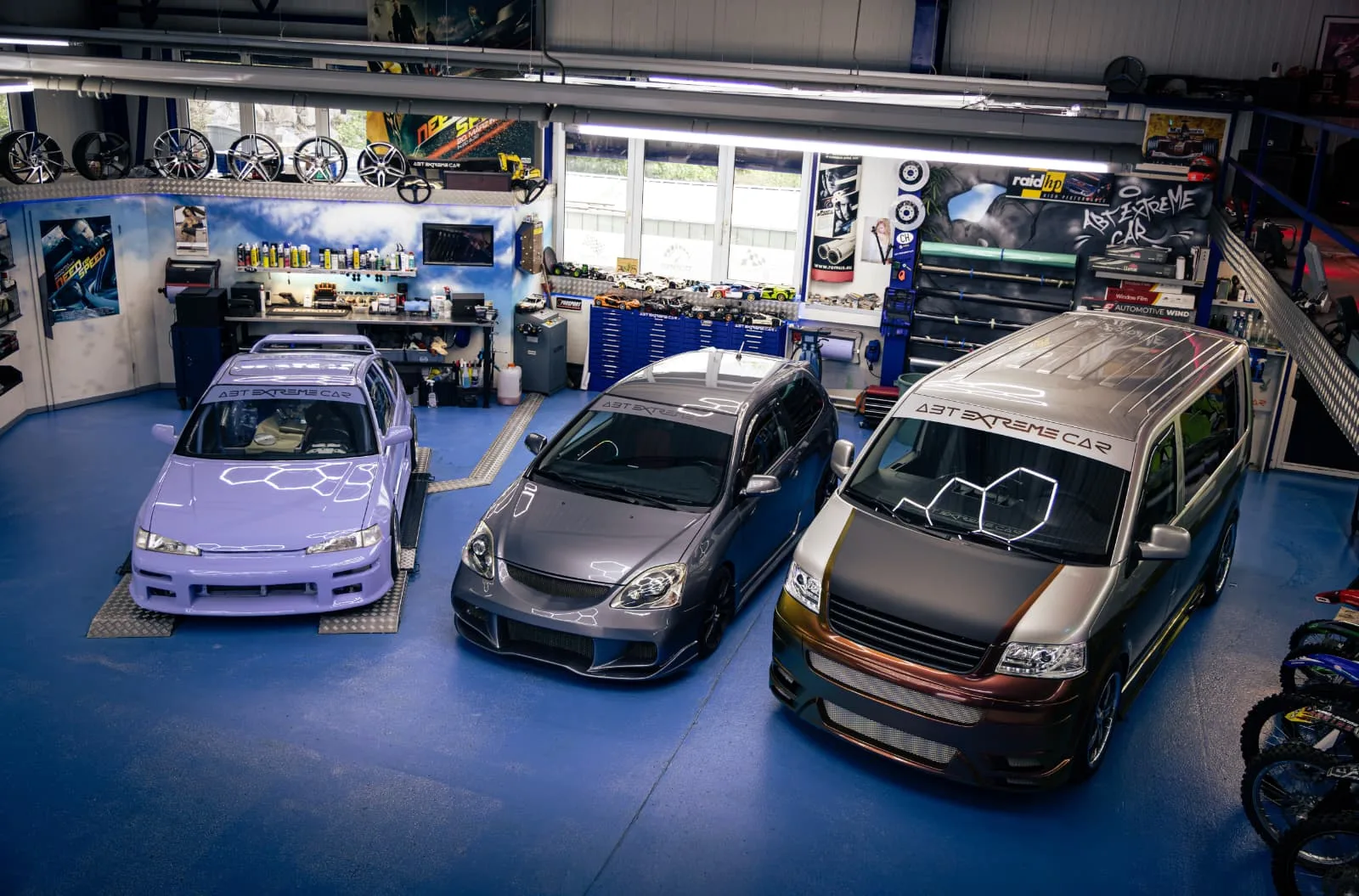 A professional tuning workshop with blue floors, tool stations, alloy wheels on display, and branded signage.