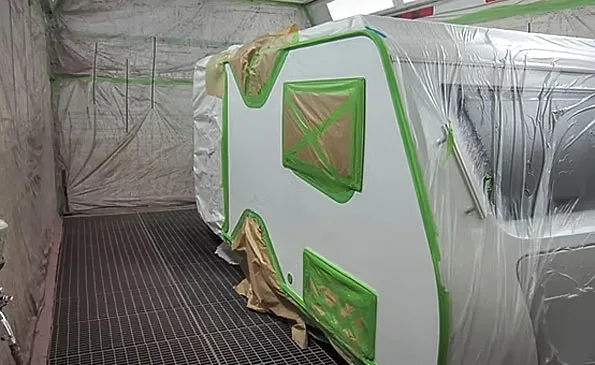 A green-and-white caravan masked and prepared for repainting inside a spray booth.