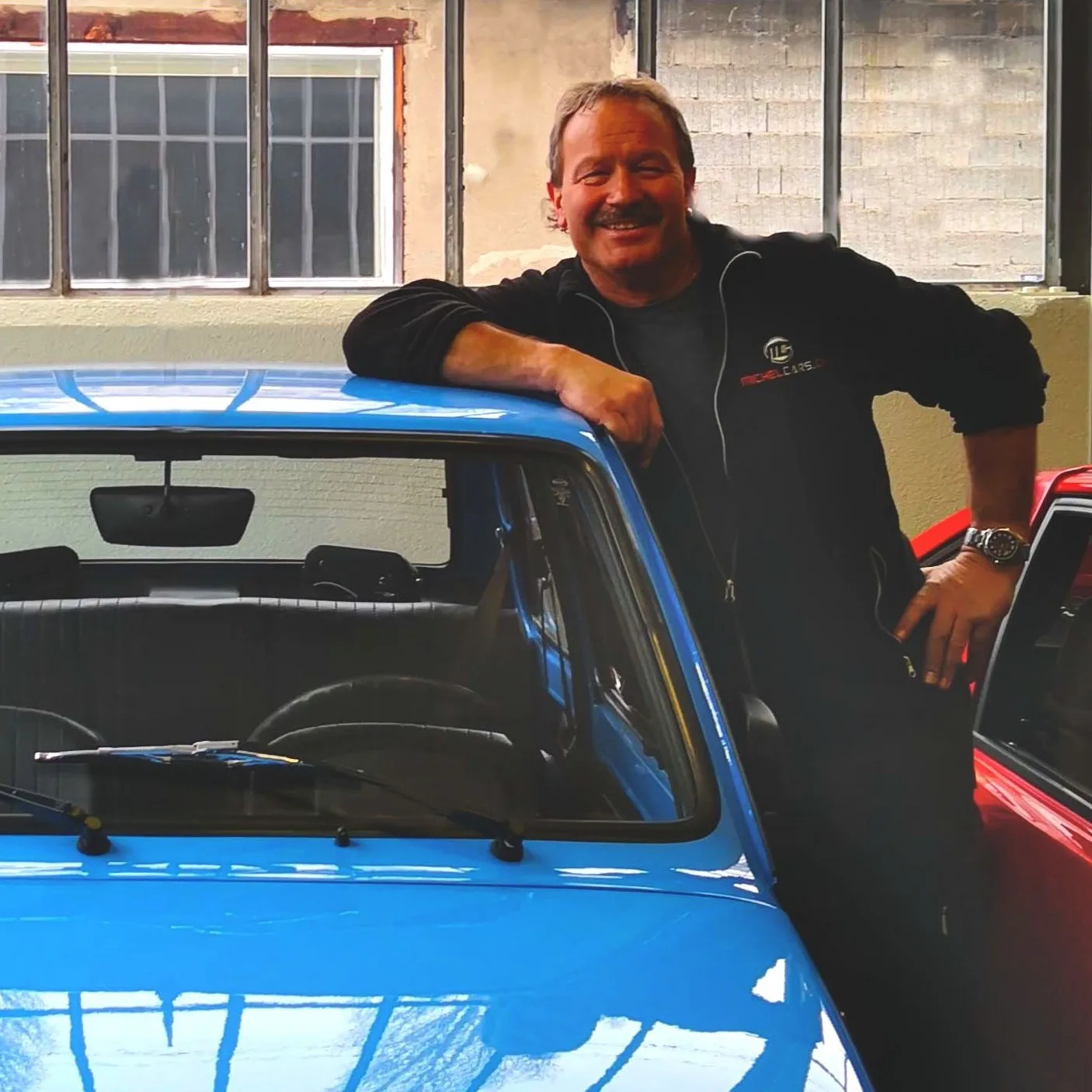 Smiling owner Paul Michel leans proudly on a classic blue car inside his garage workshop.