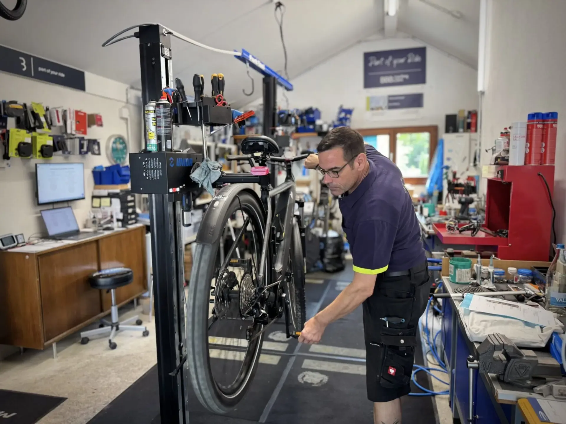 Mechanic bends over a bicycle on a Unior repair stand, carefully working on the rear wheel.
