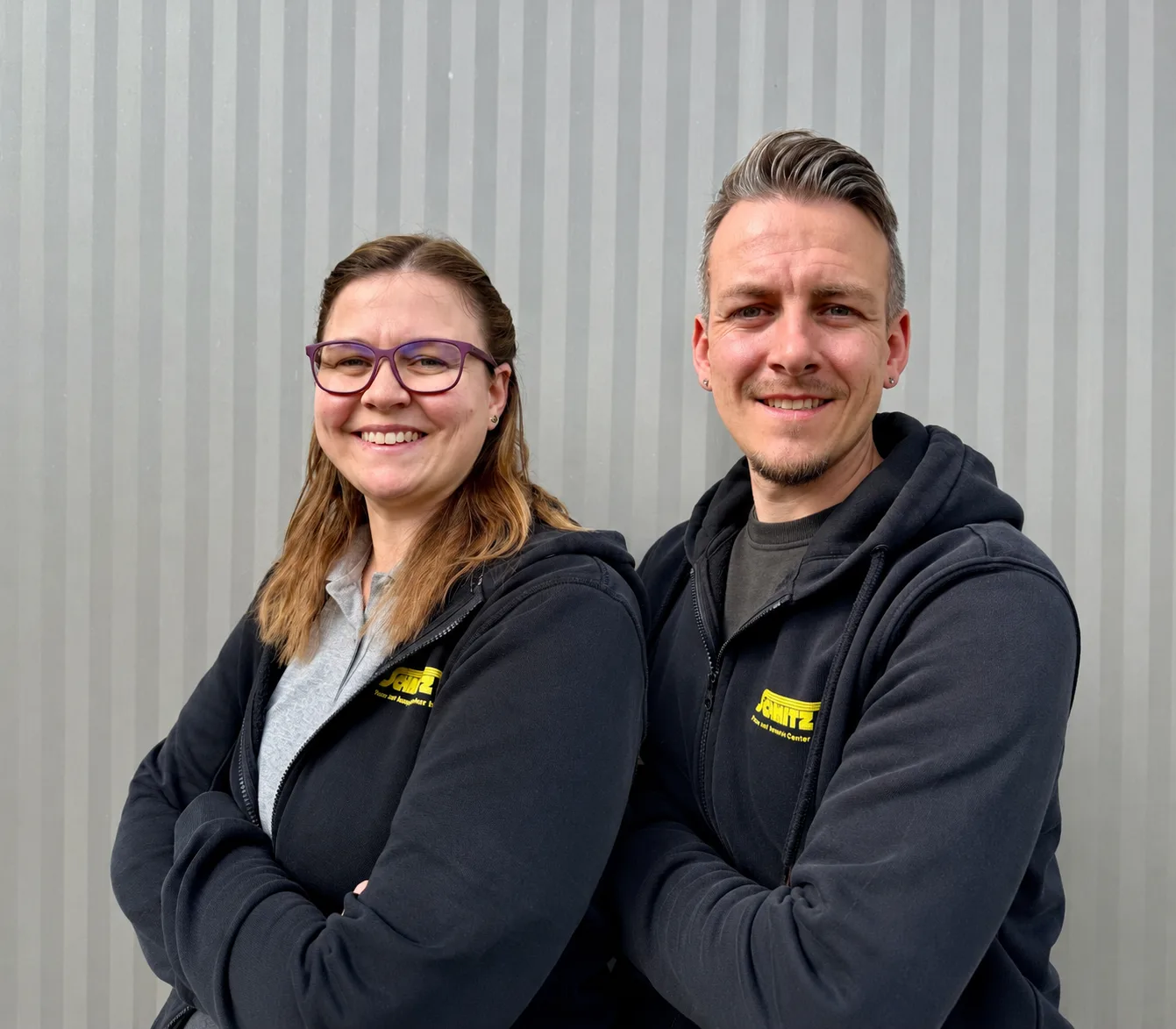 Two smiling Schmitz AG employees in branded hoodies pose confidently in front of a corrugated metal wall.